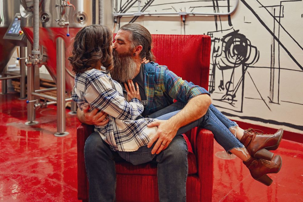 Couple sharing a kiss while sitting in a red velvet chair inside a brewery with mural wall art.