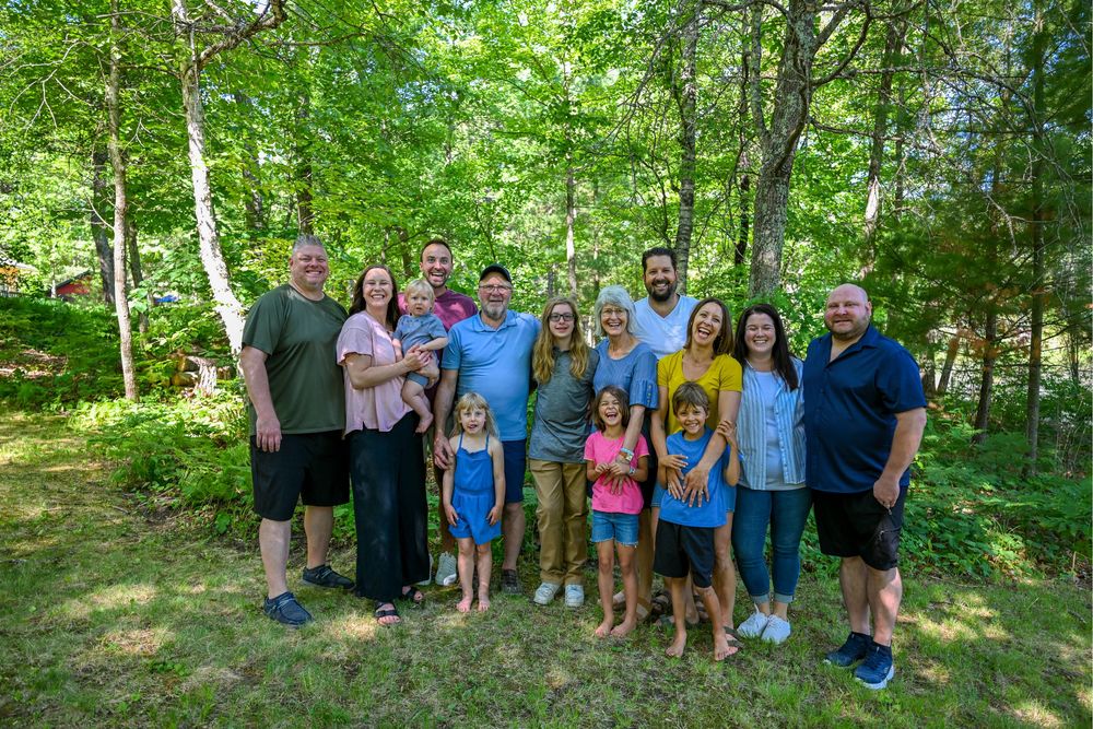 Large family group smiling together outdoors in a lush green wooded setting on a sunny summer day.