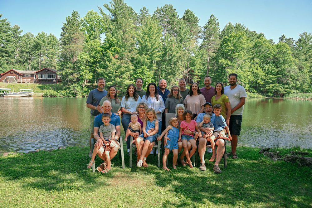 Large family group photo outdoors by a lake, surrounded by lush green trees on a sunny summer day.