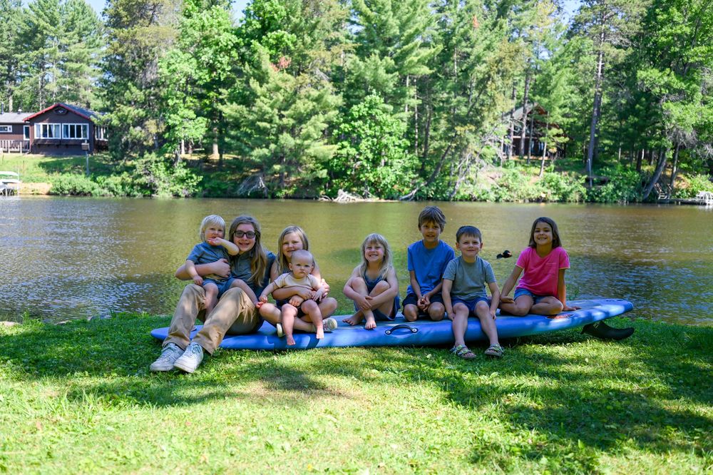 Group of children sitting on a blue mat by a peaceful lake surrounded by lush green trees on a sunny day.