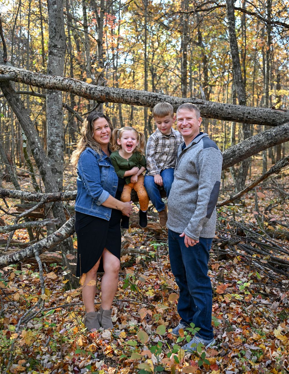 Smiling family of four in casual clothes posing together in an autumn forest with fallen leaves and a large log behind them.
