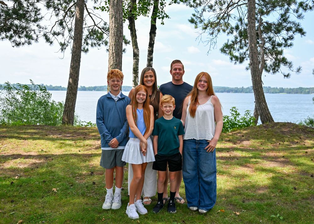 Family of six smiling outdoors near a lake surrounded by tall pine trees on a sunny day.