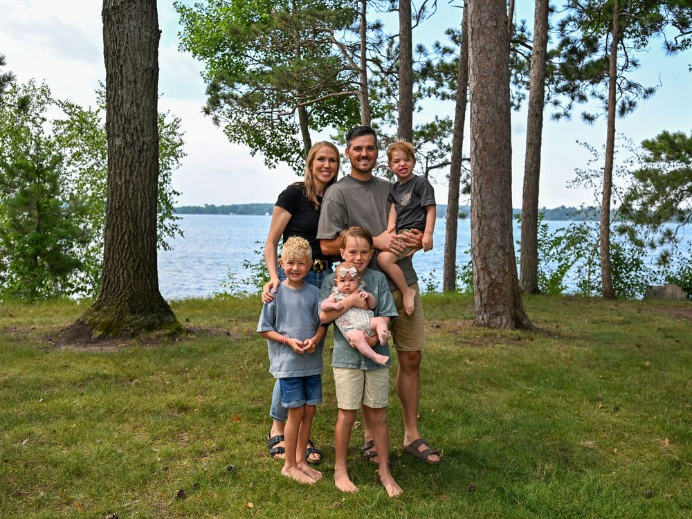 Family of six posing outdoors near a lake surrounded by tall pine trees on a sunny summer day.