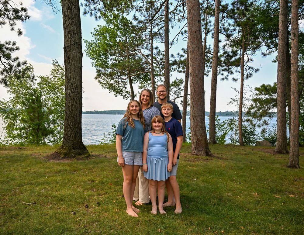 Family of five smiling outdoors near a lake surrounded by tall pine trees on a lush green lawn.
