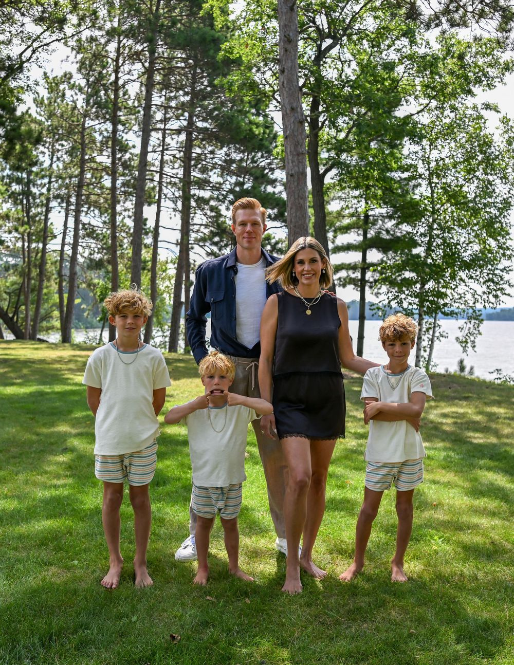 Family of five posing outdoors on green grass surrounded by tall pine trees near a lake on a sunny day.