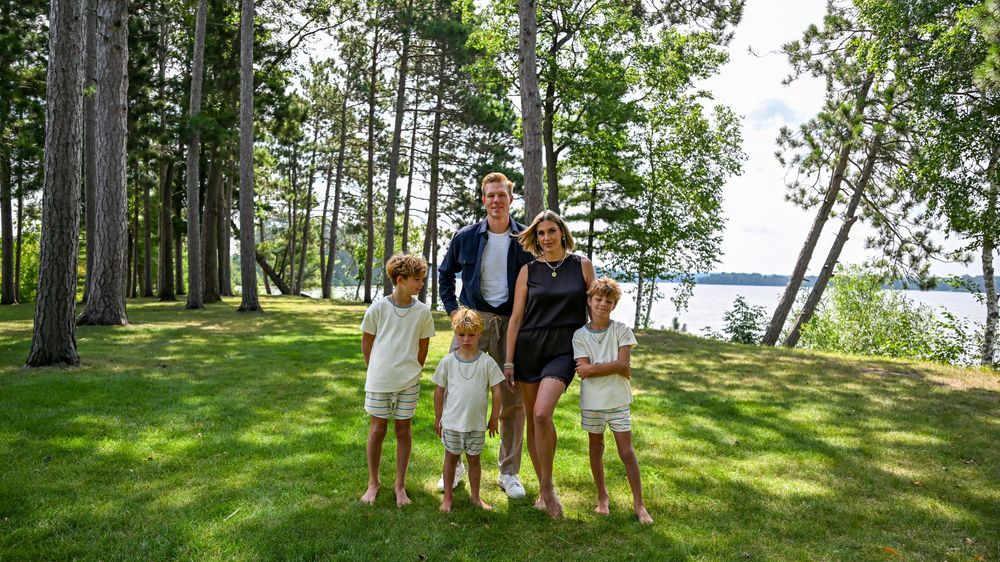 Family of five posing outdoors on lush green grass surrounded by tall pine trees near a lake on a sunny day.