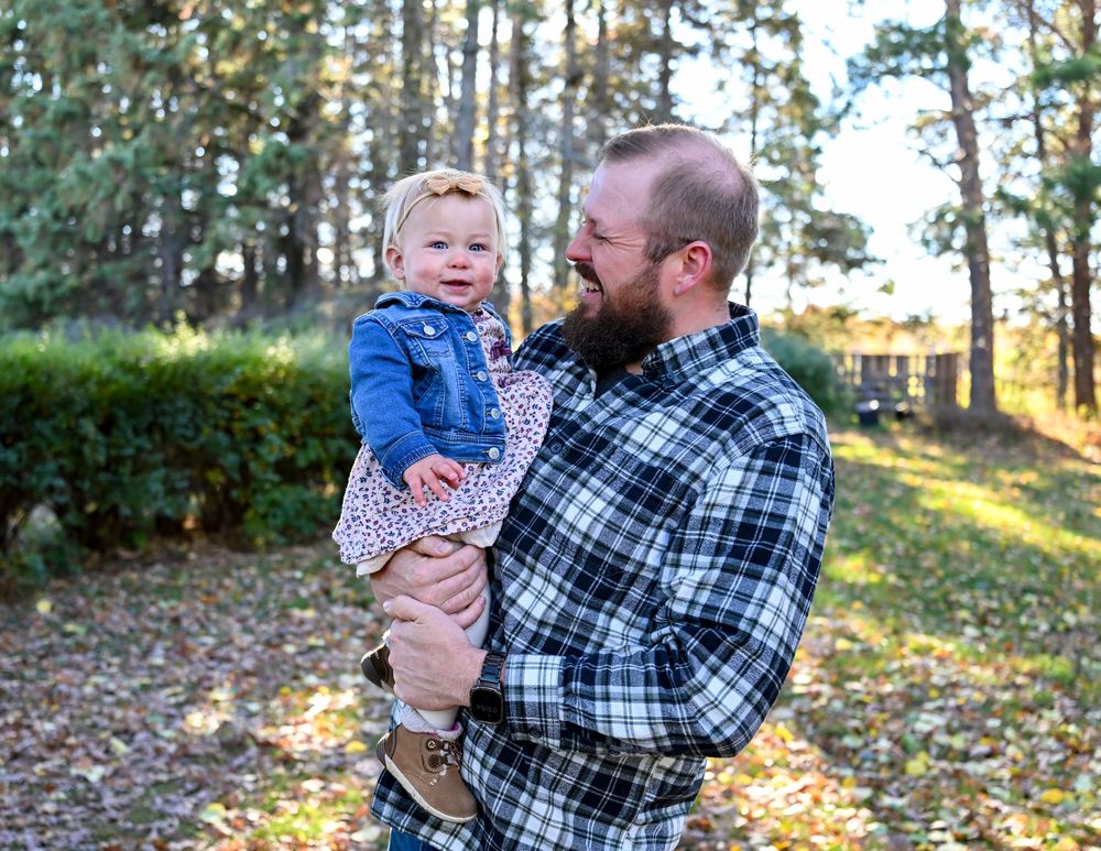 Smiling bearded man in a blue plaid flannel shirt holds a happy toddler in a denim jacket and floral dress outdoors among sunlit pine trees.