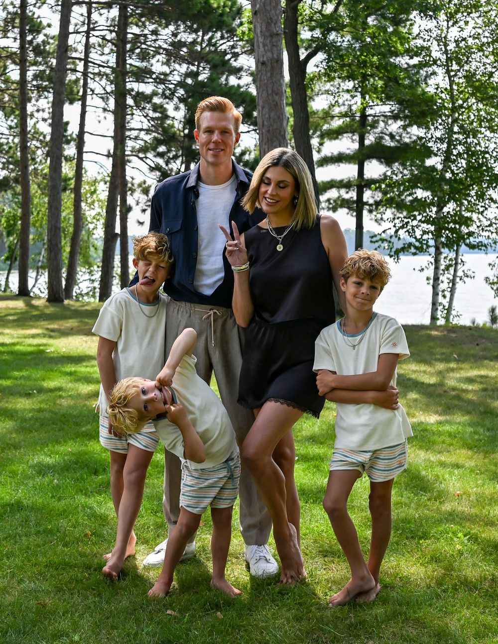 Family of five posing outdoors on green grass surrounded by tall pine trees near a lake on a sunny day.
