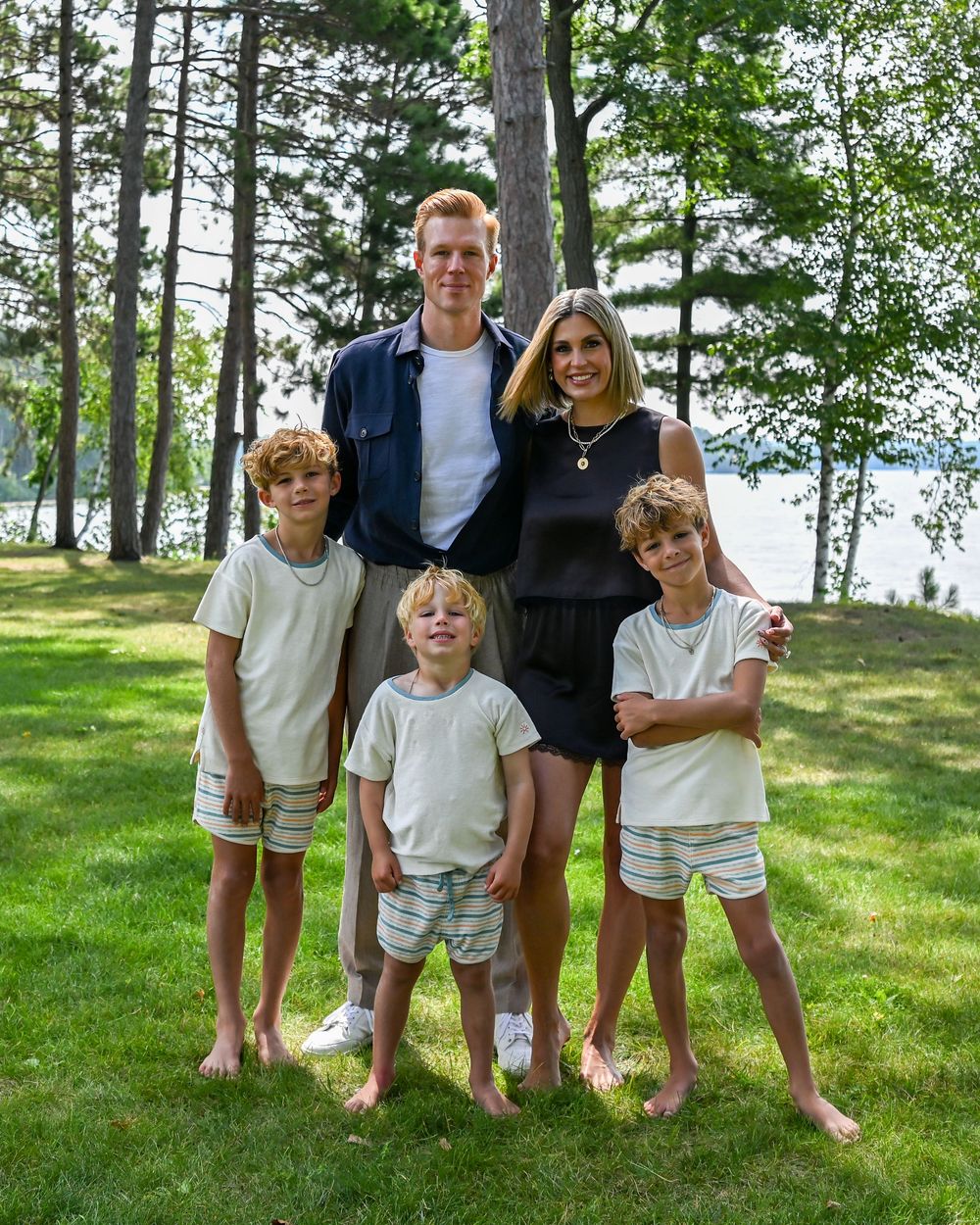 Family of five posing outdoors on green grass surrounded by trees near a lake on a sunny summer day.