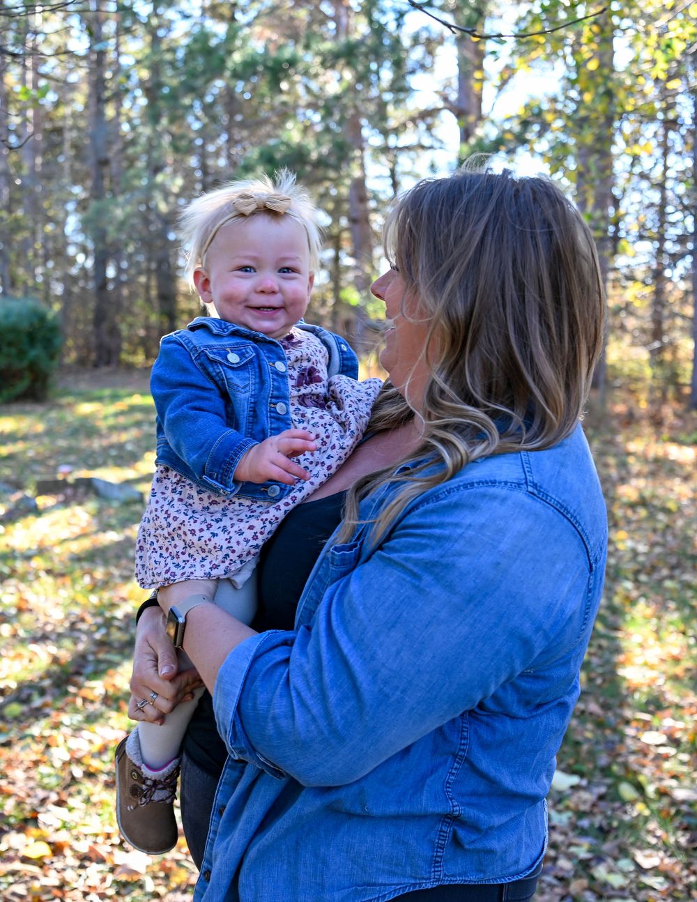 A smiling baby girl in a denim jacket and floral dress is held by a woman in a blue shirt outdoors among sunlit autumn trees.