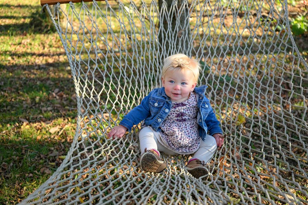 Smiling toddler in a denim jacket and floral top sits on a rope net hammock outdoors on a sunny, leaf-strewn lawn.