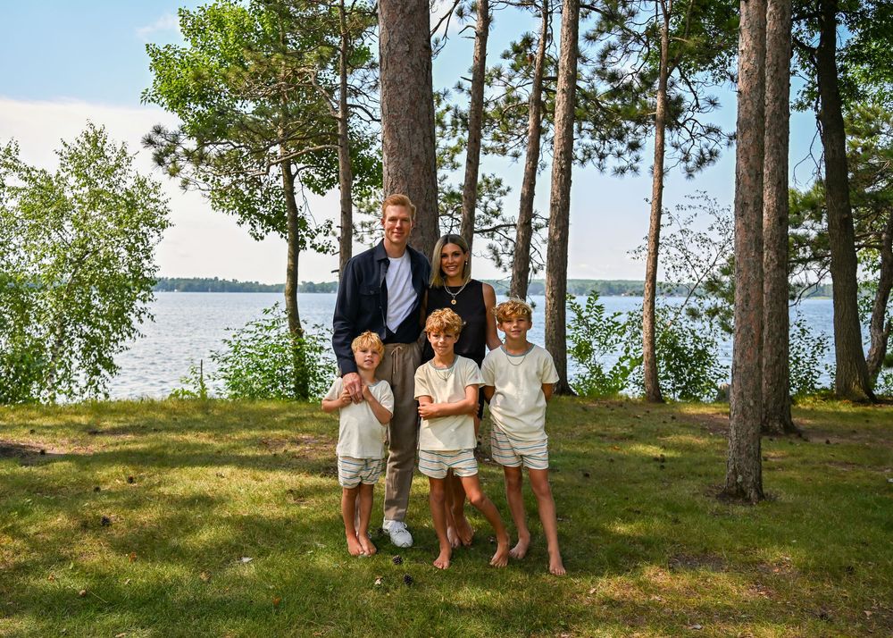 Family of five posing outdoors near a lakeside with tall pine trees on a sunny day.
