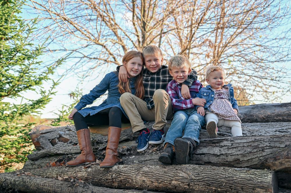 Four smiling siblings in casual fall clothes sit close together on a stacked log pile with bare trees and blue sky behind them.