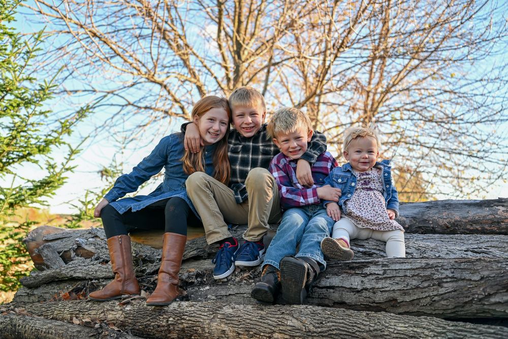 Four smiling children sit together on a stack of weathered logs outdoors beneath bare trees and a clear blue sky.