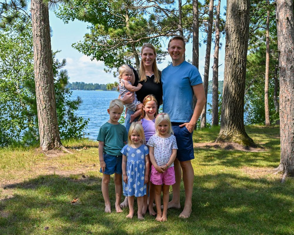 Family of seven posing outdoors near a lake surrounded by tall pine trees on a sunny summer day.