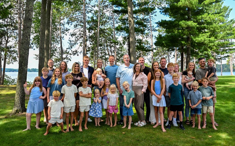Large multigenerational family group portrait outdoors near lake, surrounded by tall pine trees on green lawn.