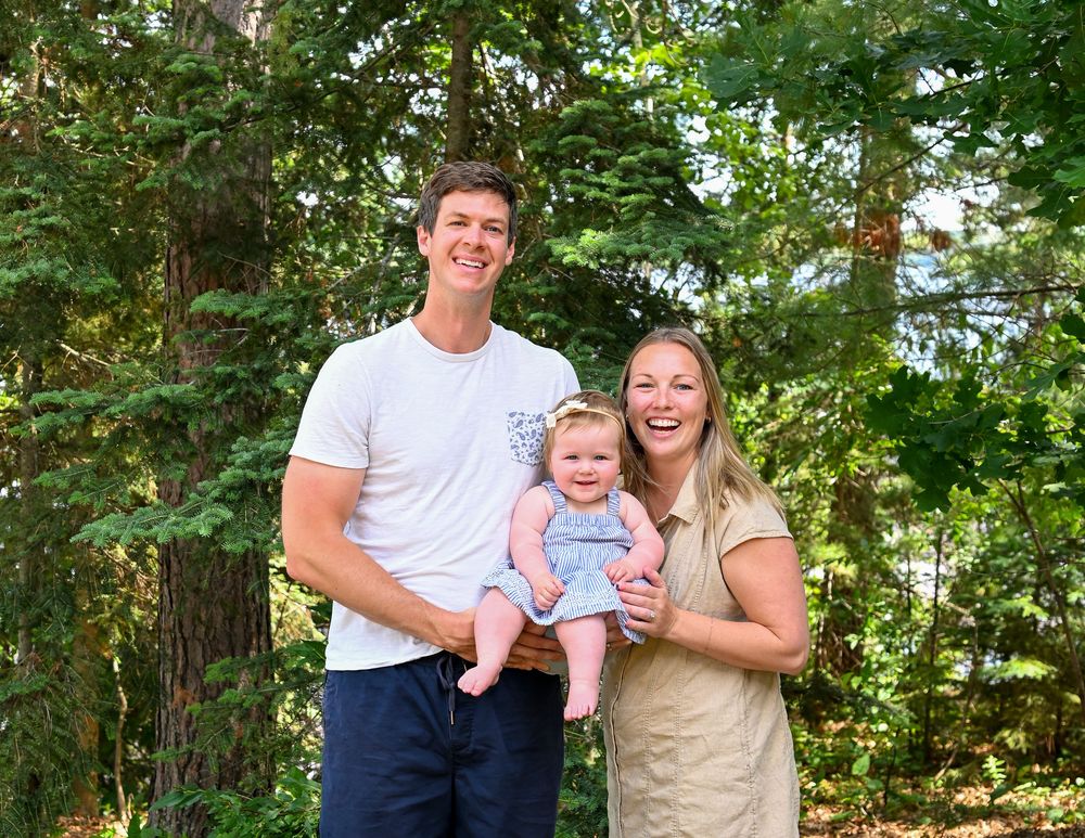 Happy young family with a baby smiling outdoors surrounded by lush green trees in a forest setting.