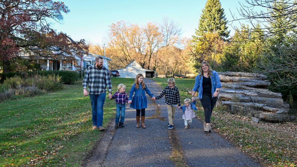 Family of six holding hands and walking down a paved path through a park lined with autumn trees and stacked logs.