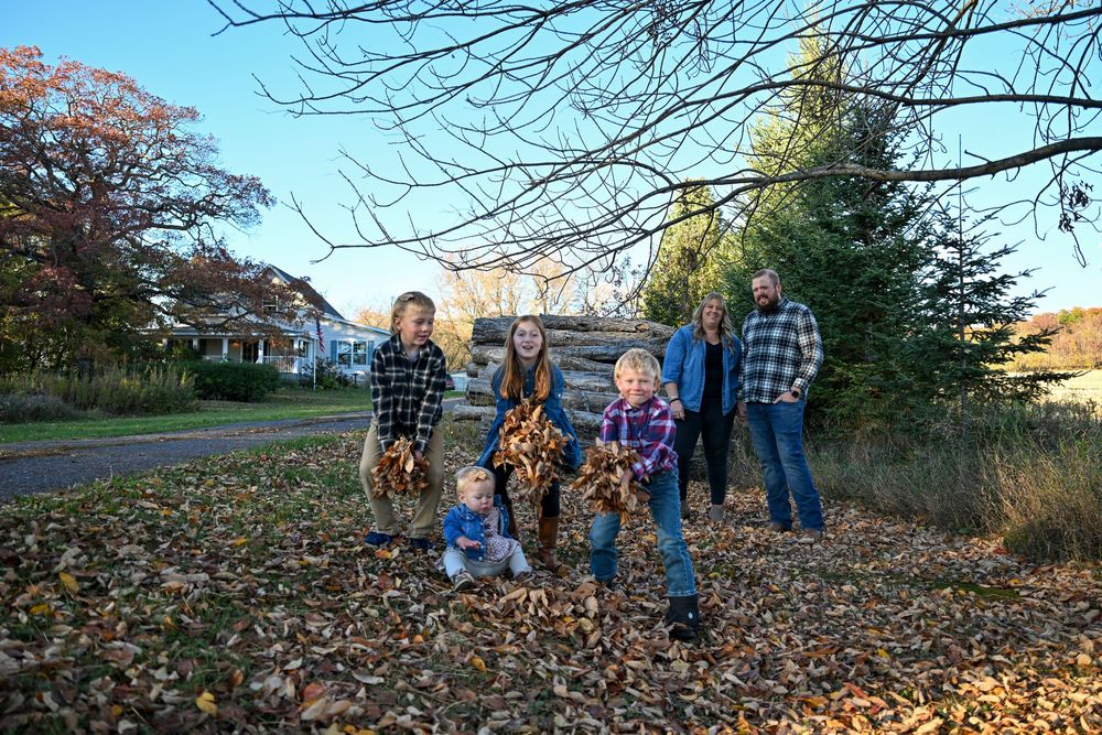 Family of six in plaid shirts tosses armfuls of colorful fallen leaves in a sunny autumn yard with bare trees and evergreens.