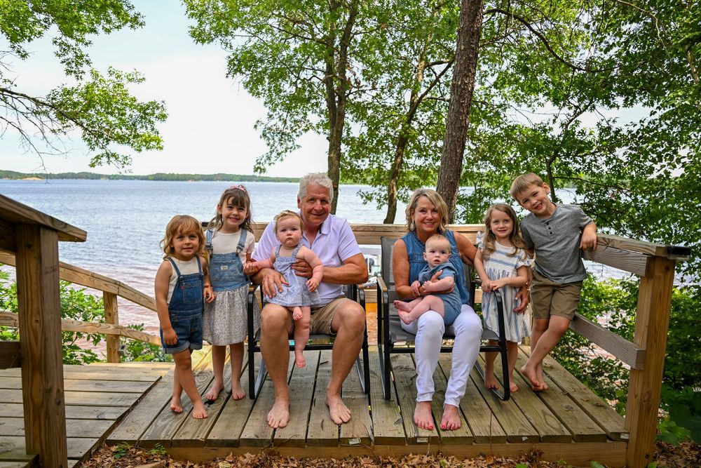 Grandparents sit on a wooden deck with six grandchildren, surrounded by lush trees and a serene lake backdrop.