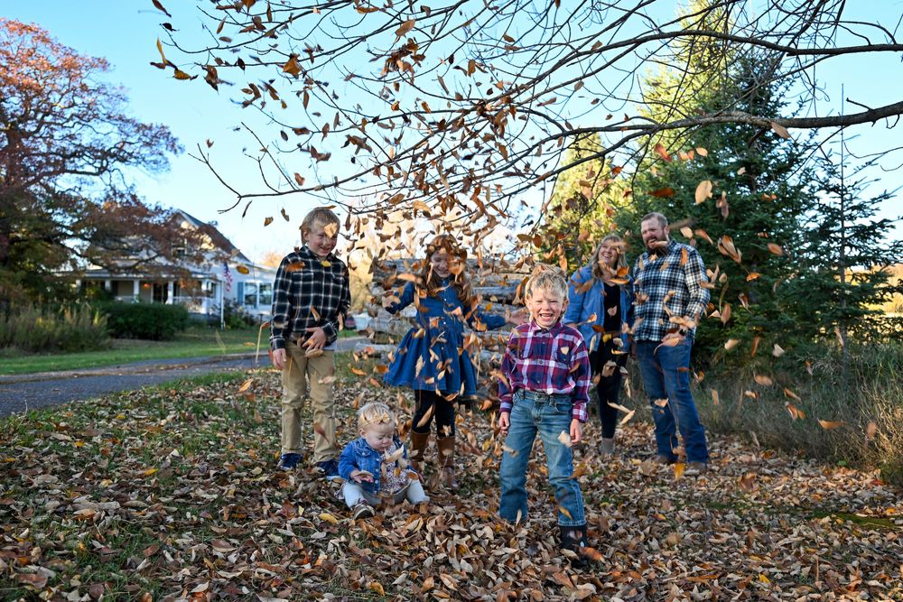 Family of adults and children in plaid and denim playing in a yard as autumn leaves fall around them on a sunny fall day.