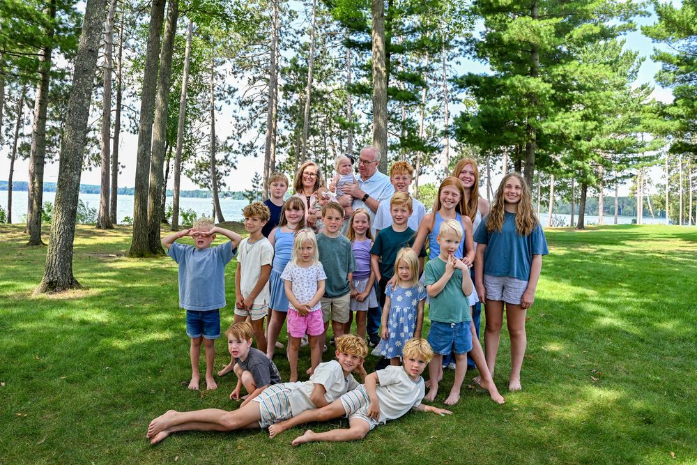 Large family group photo with adults and children posing outdoors on green lawn surrounded by tall pine trees near a lake.