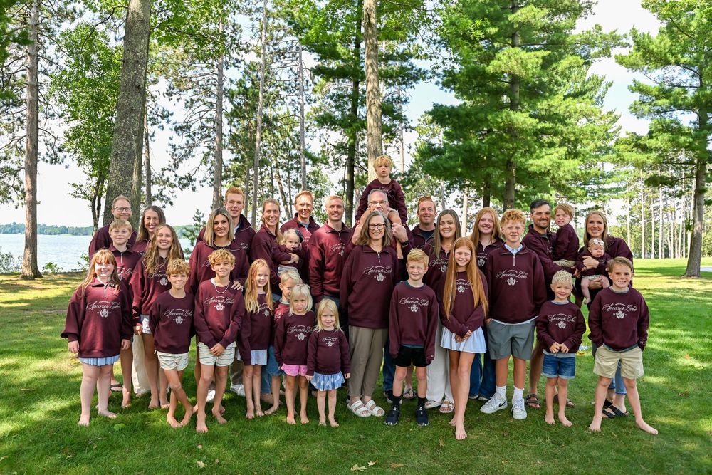 Large family group photo outdoors in matching maroon shirts, posing on green lawn with trees and lake in background.