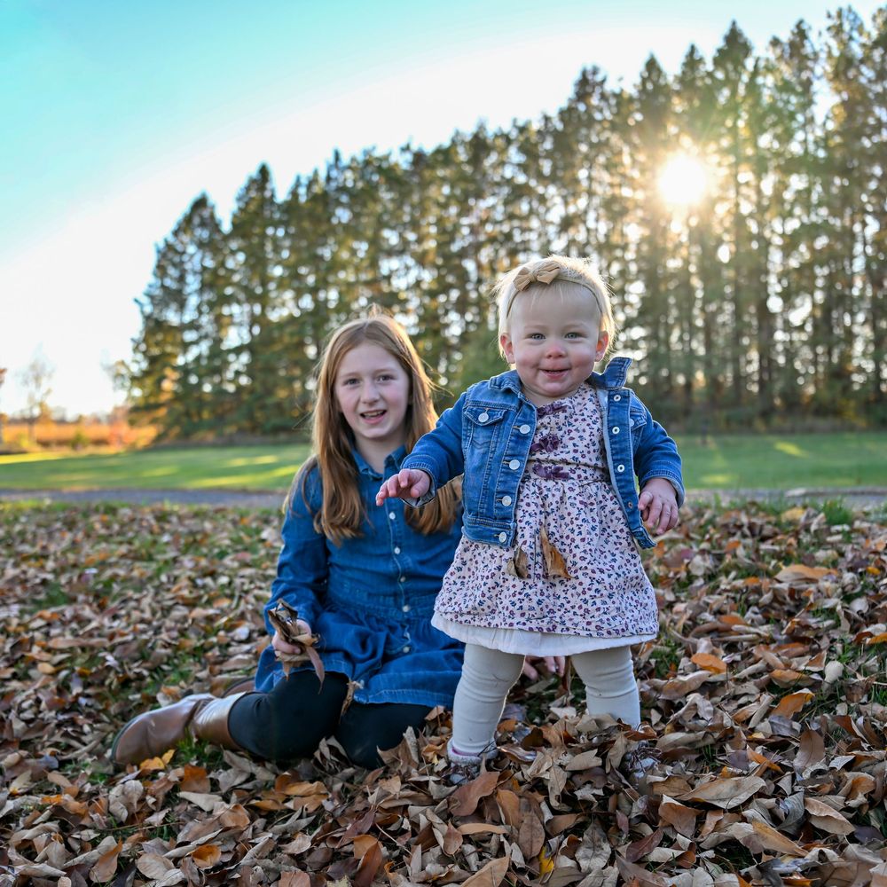 Two young sisters in denim jackets play in a pile of fallen autumn leaves with tall evergreen trees and golden sunset light behind them.