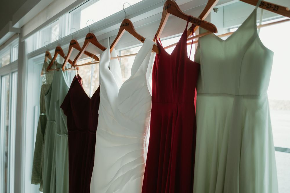 Colorful bridesmaid dresses in sage, burgundy, and white hanging on wooden hangers by a bright window.