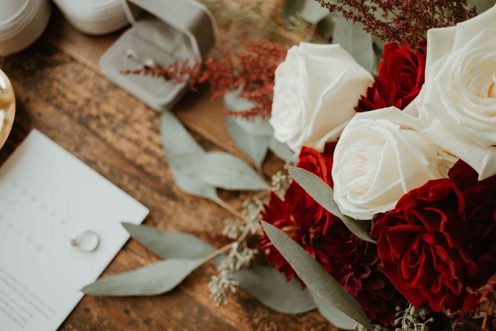 Bridal flat lay featuring white and red rose bouquet, wedding invitation, rings, and eucalyptus on rustic wood.