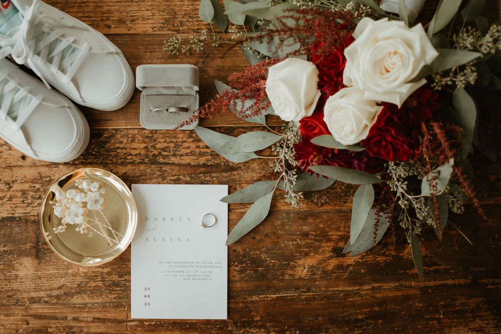 Wedding detail flat lay with bouquet, white sneakers, ring box, invitation, and accessories on rustic wood table.