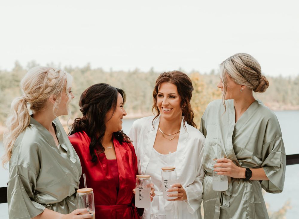 Bride in white robe laughing with three bridesmaids in satin robes on an outdoor balcony with scenic water views.