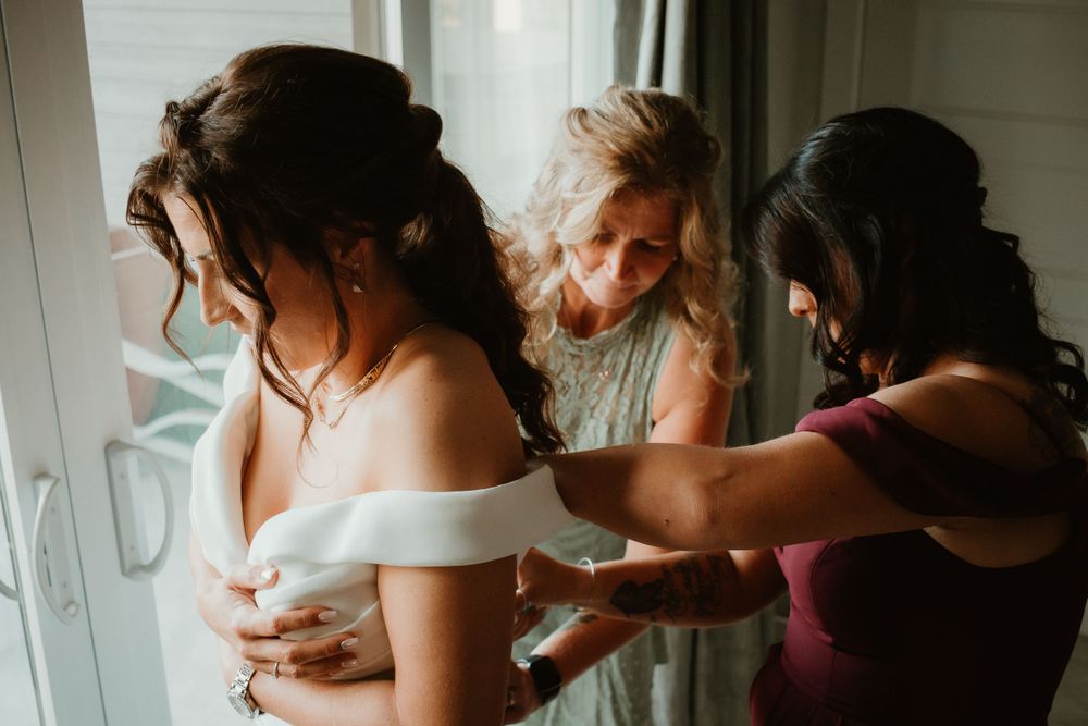 Two bridesmaids help a bride adjust her off-shoulder white wedding gown near a bright window.