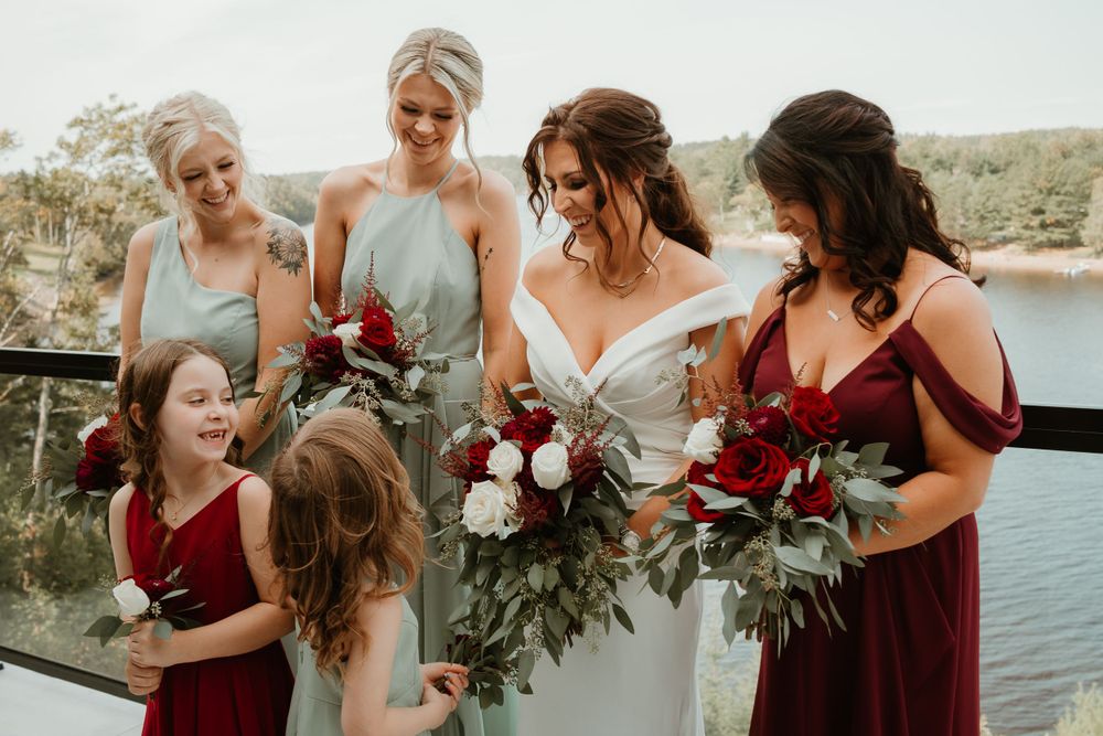 Bride in white gown with bridesmaids in sage and burgundy dresses holding red and white bouquets by a lake.