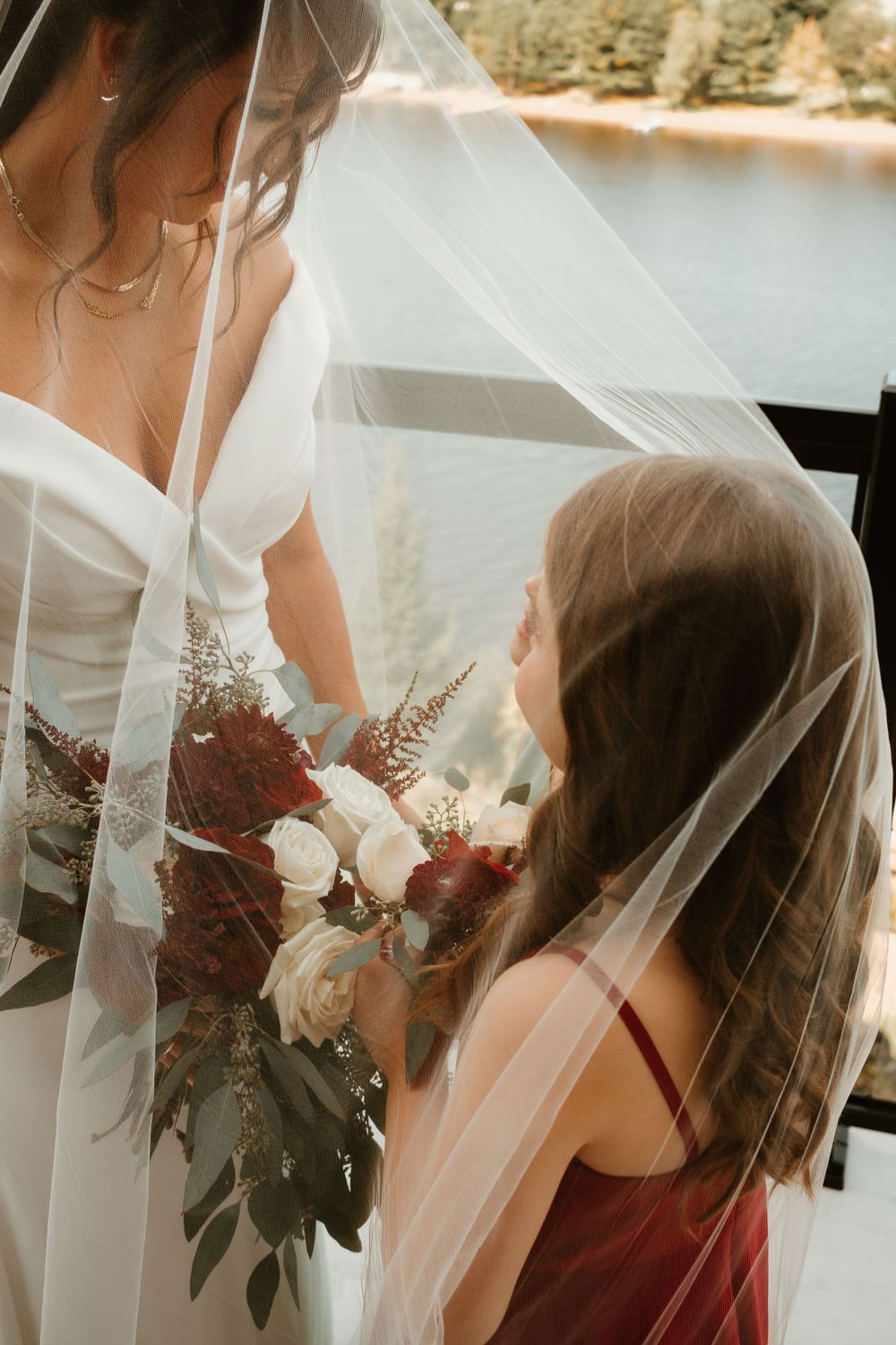 Young flower girl in burgundy dress admires bride's bouquet of red and white roses under a flowing veil.