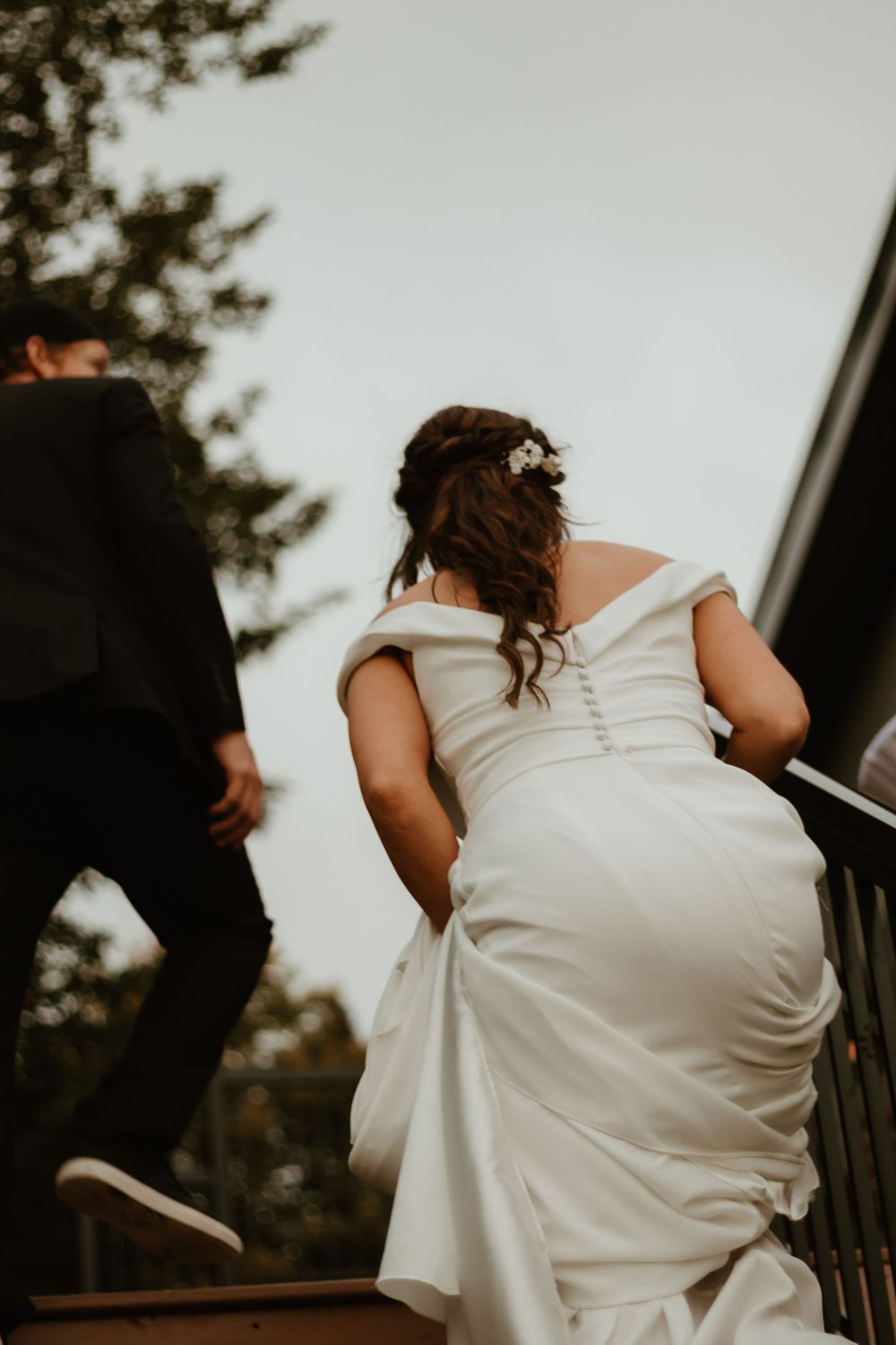 Bride in elegant off-shoulder white wedding gown walks up stairs holding dress train.