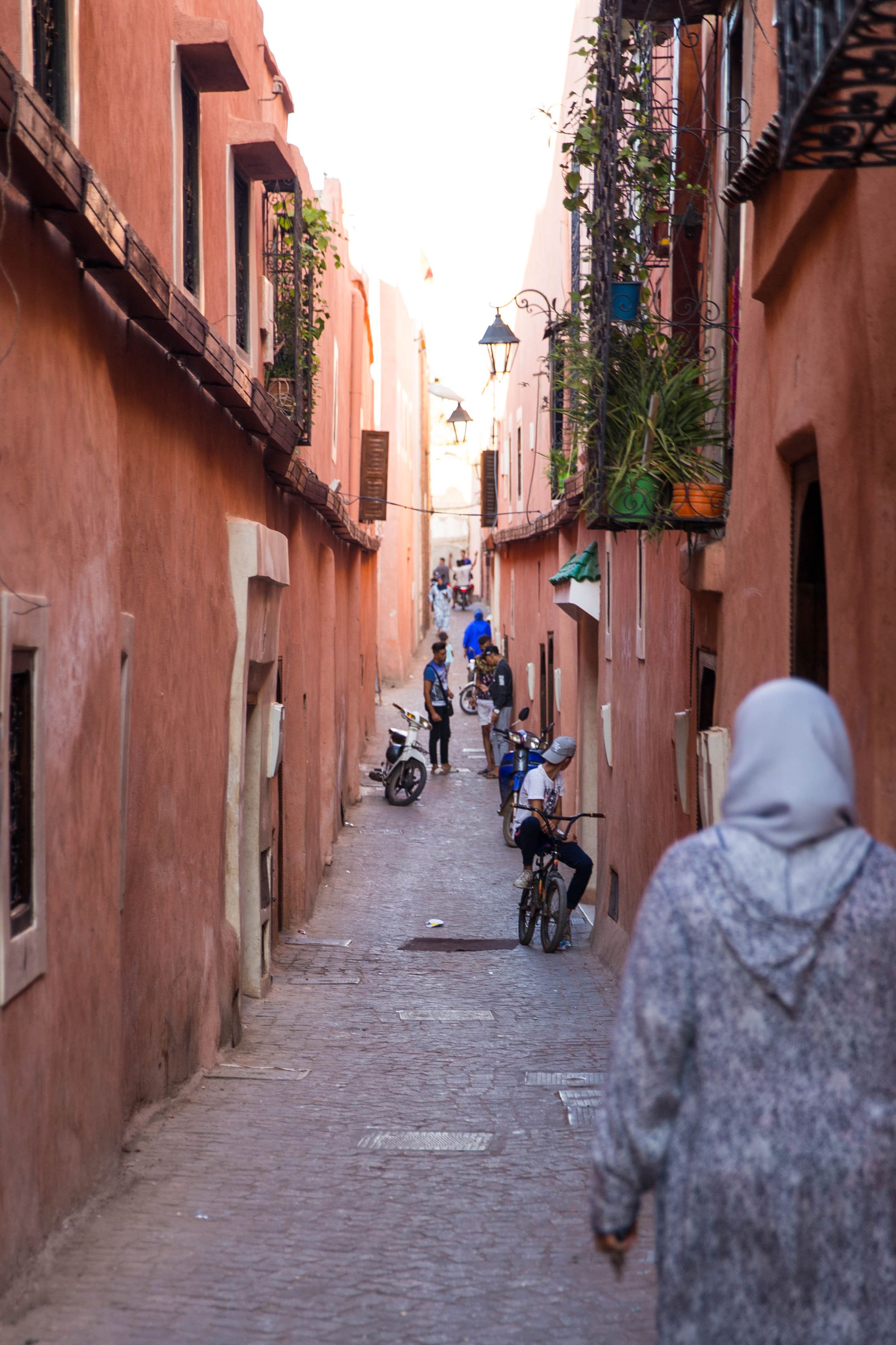 Narrow alleyway in Marrakech medina with terracotta walls, parked motorcycles and pedestrians walking.