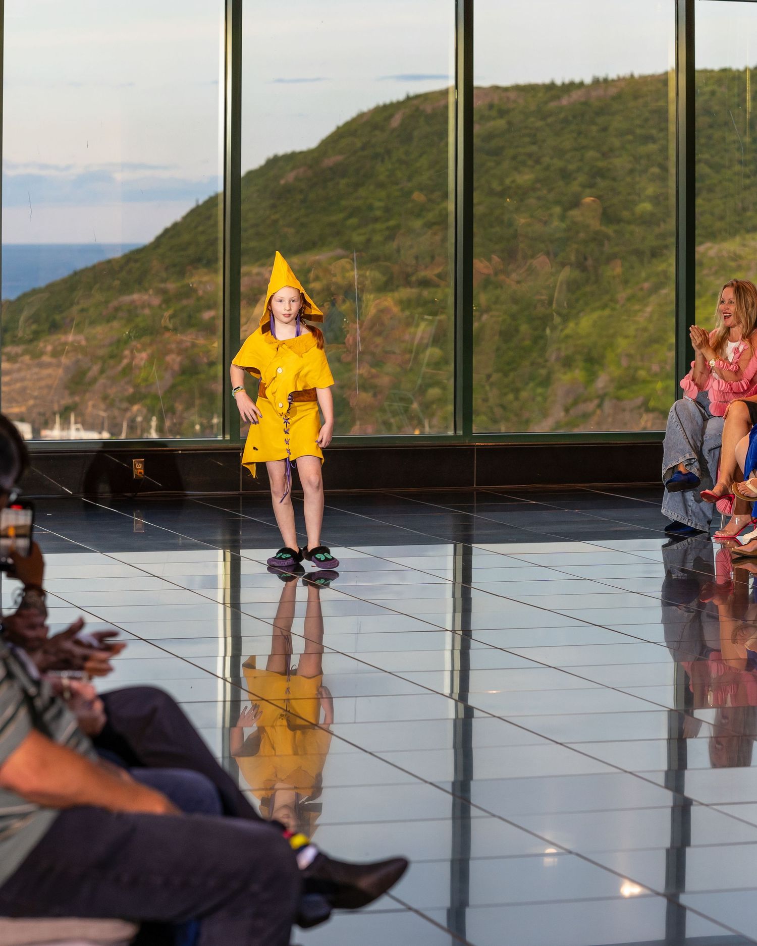 A model in a bright yellow raincoat walks the runway with mountain views visible through large windows behind.