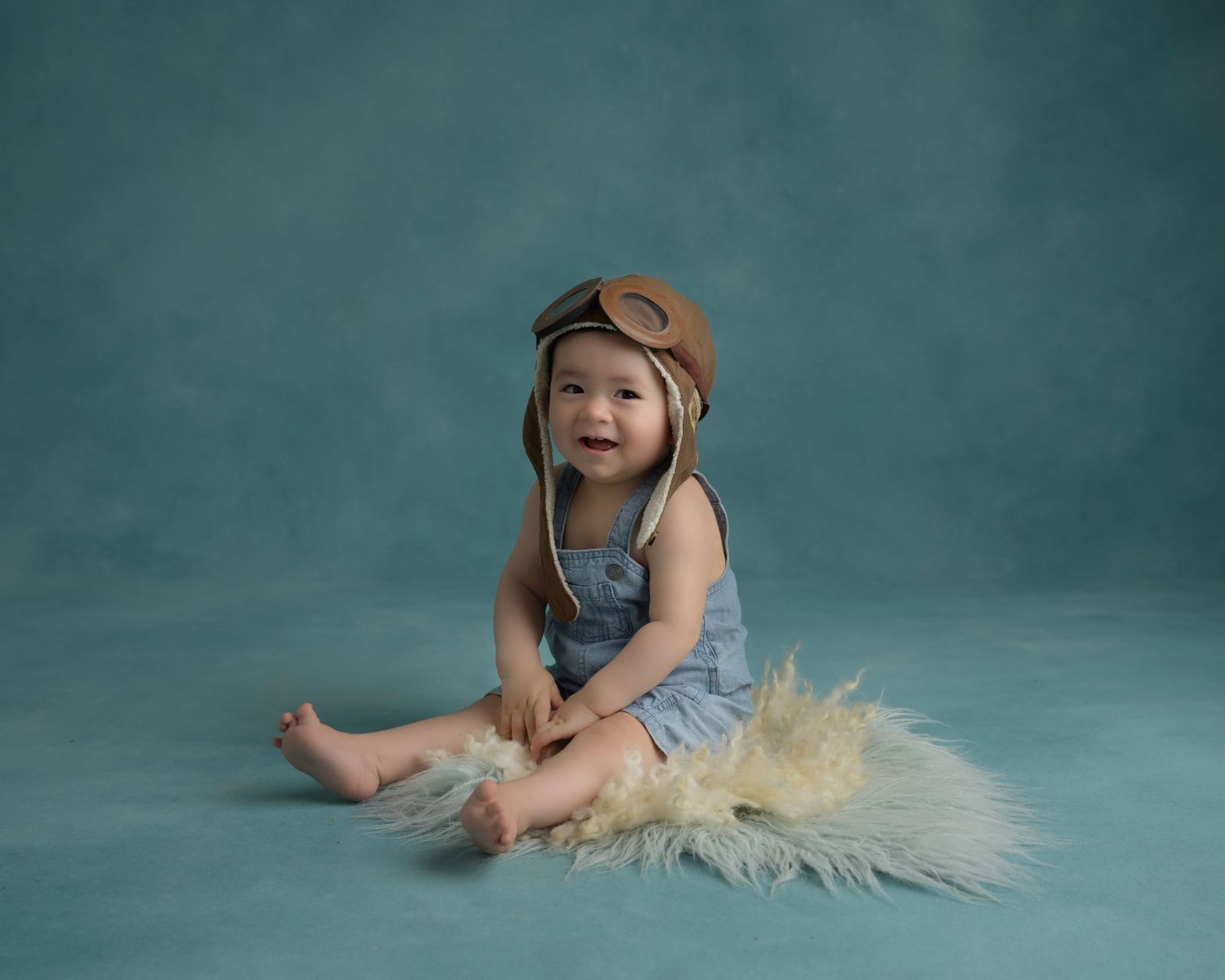 Baby wearing aviator hat and denim overalls sits on fur against teal background.