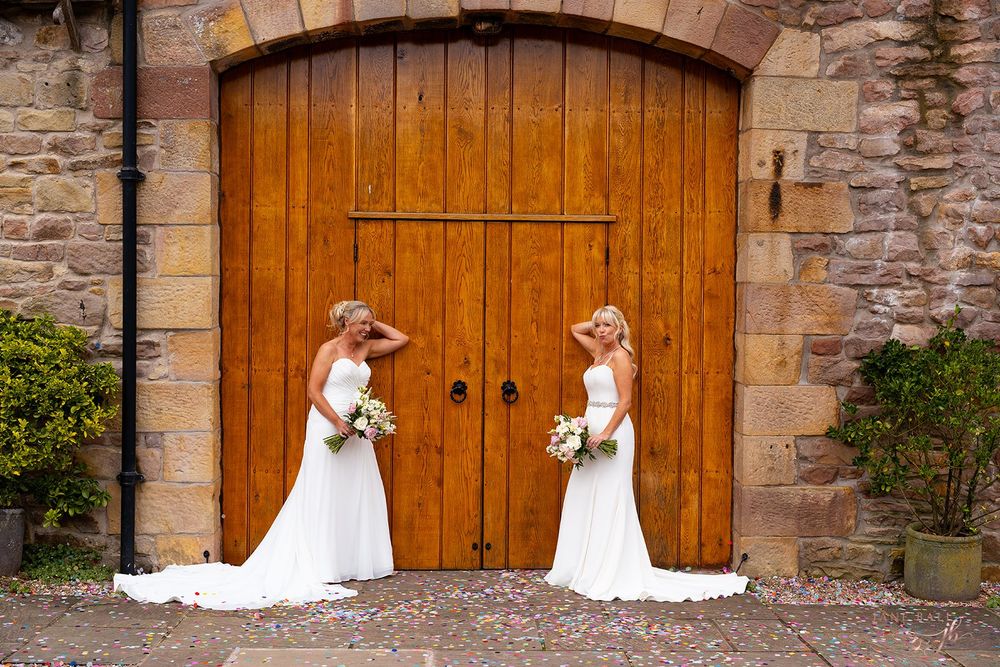 Two brides in white gowns holding bouquets stand before rustic wooden doors at a stone venue. Browesholme Hall. Jane Ball Photography