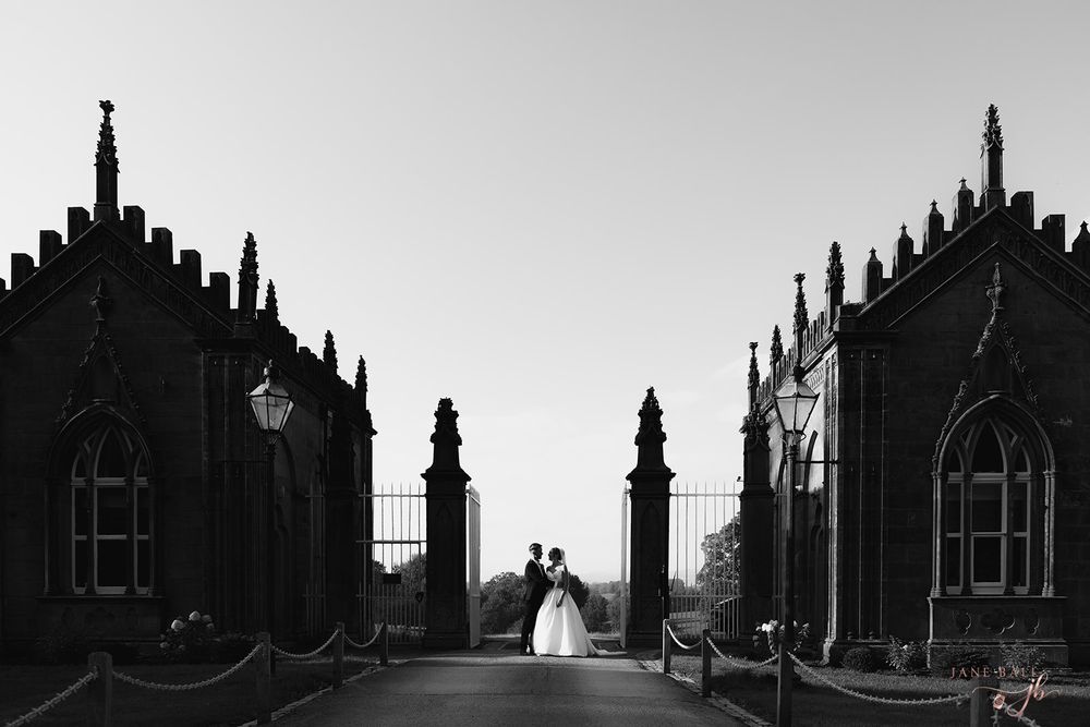Black and white wedding photo of couple kissing at ornate gothic gate entrance on a long driveway. Gisburne Park Estate. Jane Ball Photograp