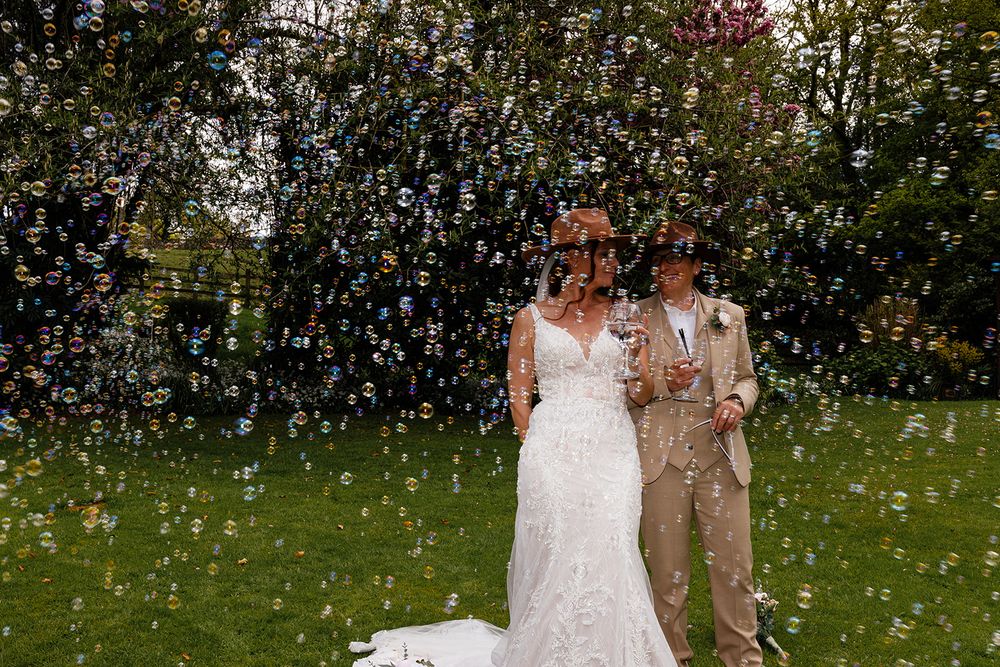 Bride and bride celebrate outdoors surrounded by floating bubbles on their wedding day in a lush green garden.