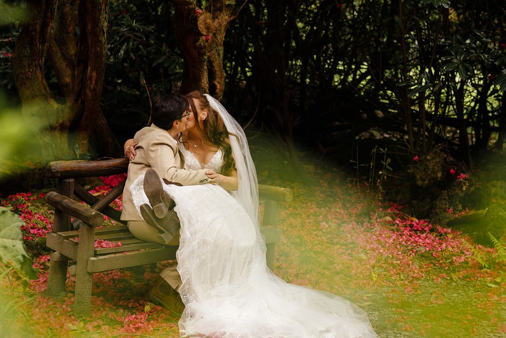 A bride in a white gown and veil kisses her wife on a wooden bench surrounded by lush greenery and red flowers.