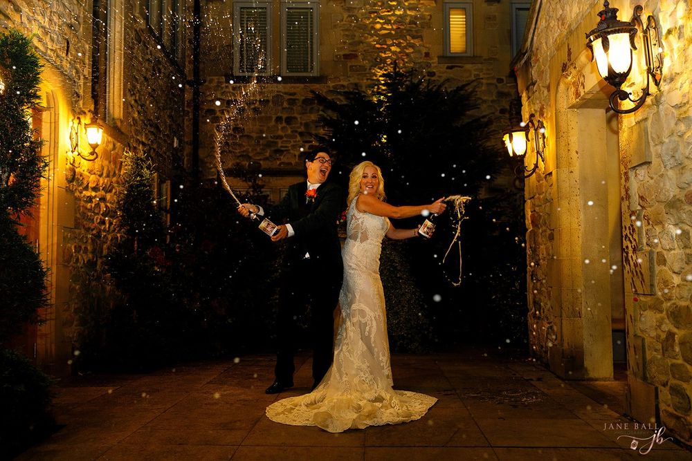 Two brides celebrate with a champagne spray at night outside stone venue in elegant wedding portrait.