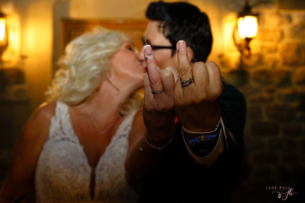 Newlyweds kissing while showing off their wedding rings to the camera in a warmly lit rustic venue.