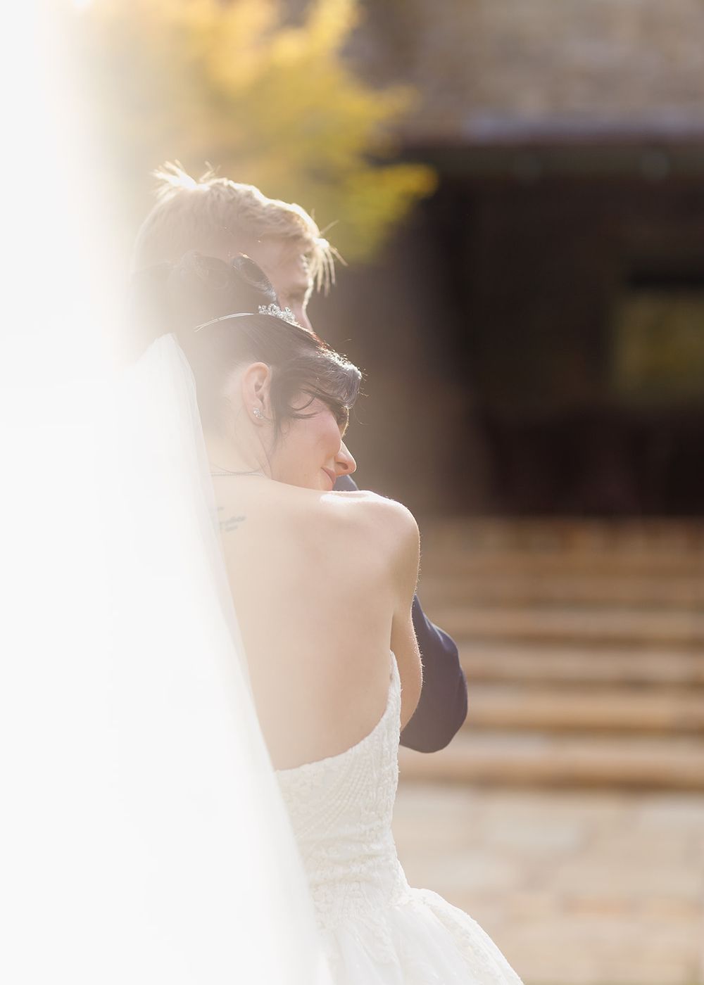 Bride in a white strapless gown embraced from behind, soft sunlight creating a dreamy bokeh background.