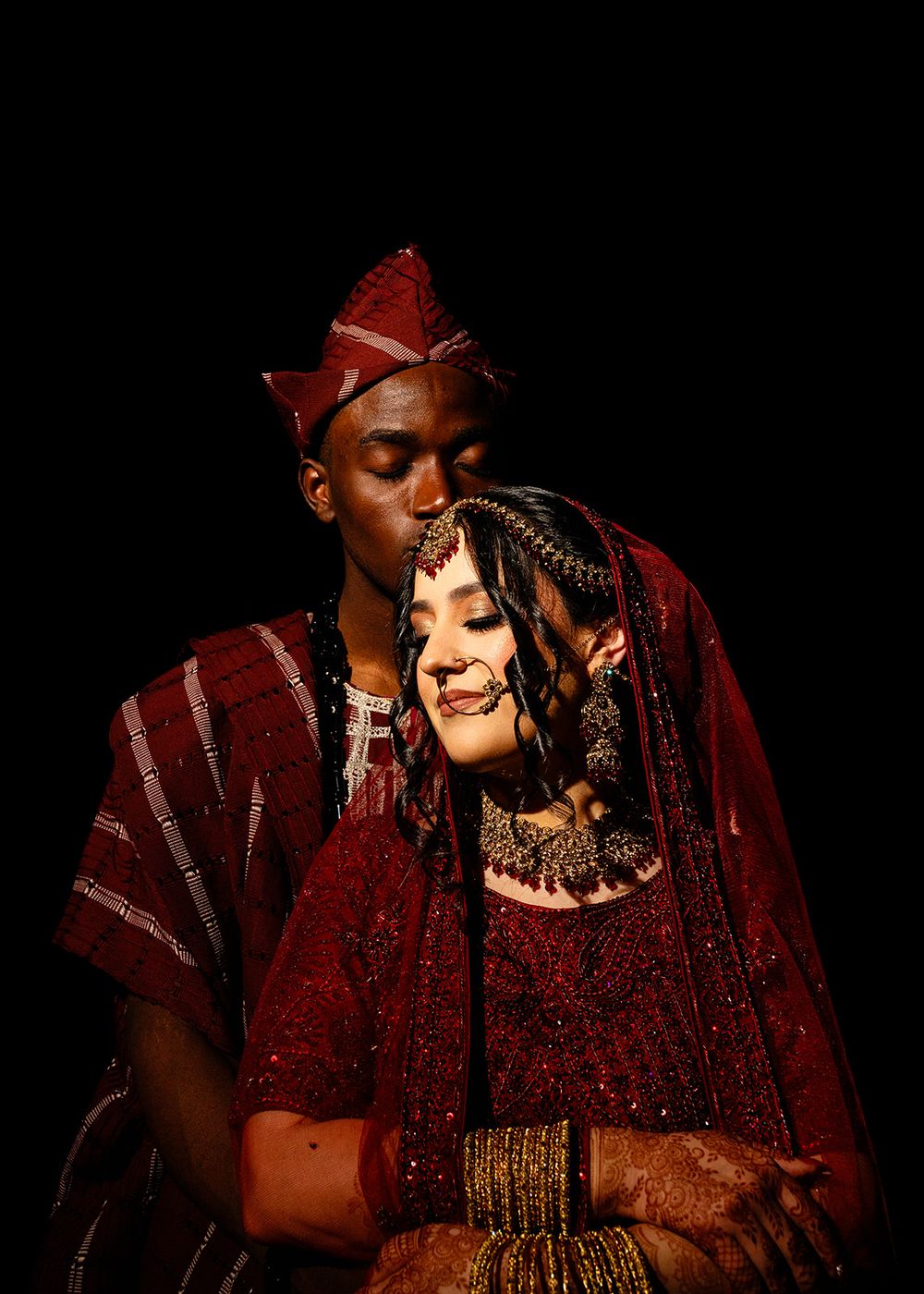 Couple in matching deep red traditional attire, woman in bridal jewelry, man in embroidered agbada and cap, dark background.