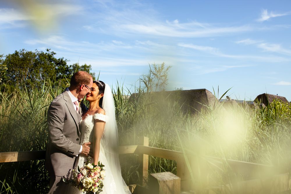 A bride and groom share a romantic kiss outdoors on a sunny day, surrounded by lush green reeds  at Brockholes Nature Reserve