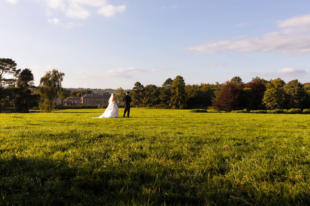 A bride and groom stand together in a vast green field with a grand estate and autumn trees in the background. Gisburne Park Estate