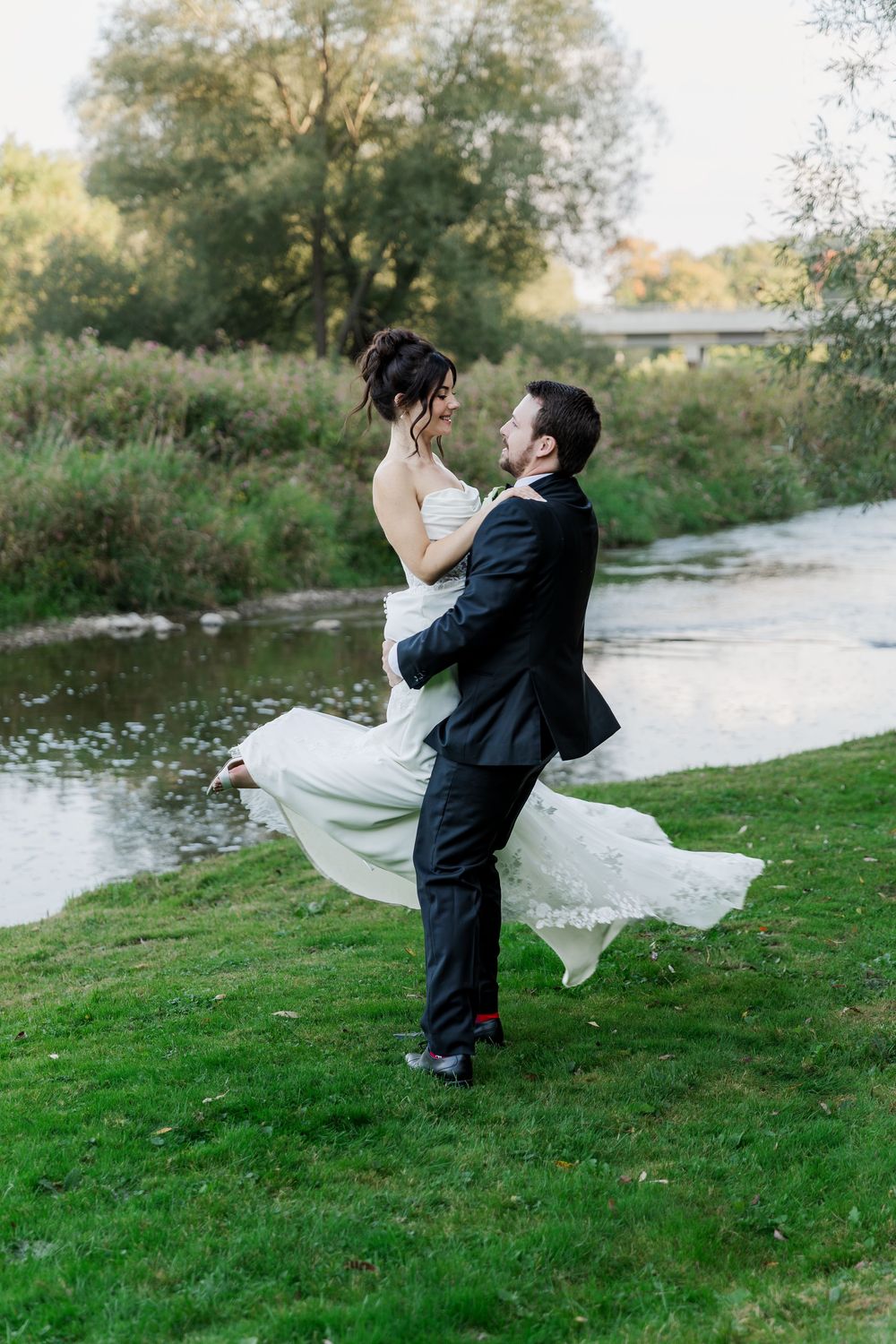 A joyful moment captured as the groom lifts his bride by a riverside on their wedding day at The Wren House in St. Jacobs, Ontario.